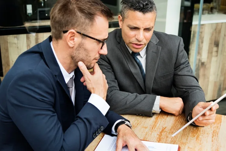 Two men in suits sitting at a wooden table, one resting his chin on his hand as they look at a document