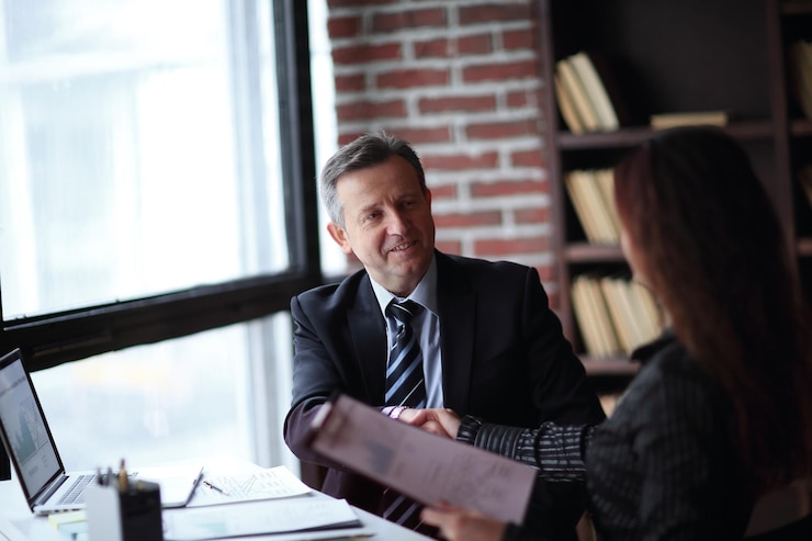 A man in a dark suit smiling at a colleague who holds a document, a laptop on the desk beside them