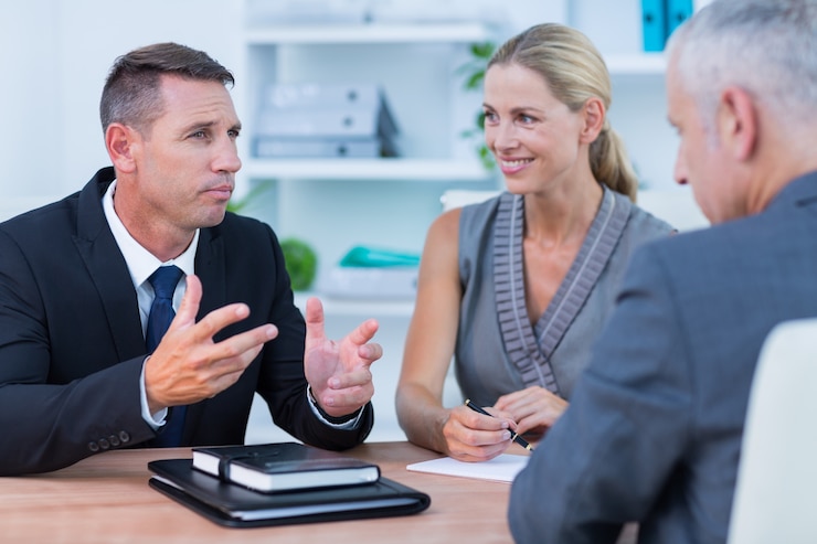 A man gesturing as he talks to a woman and a second man across a desk, with a notebook in front of him