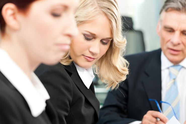 Three people in dark business clothes at a meeting table, two of them looking down at a paper document