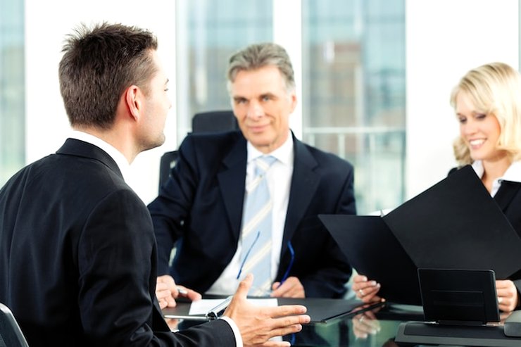 A man in a dark suit with his back to the camera speaking to two colleagues who hold a black folder