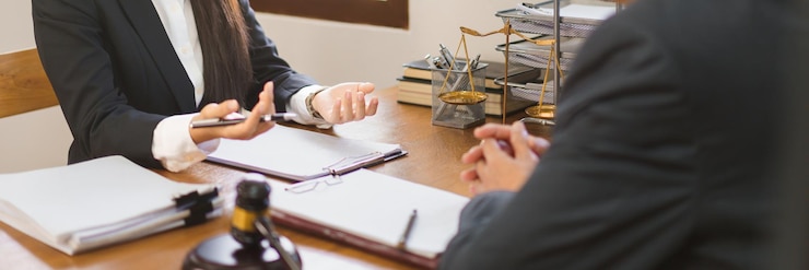 A seated woman gesturing over open documents, with a gavel and a set of scales on the desk