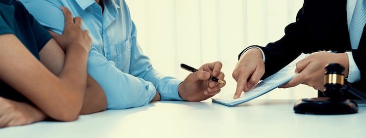 A person in a suit pointing at a document with a pen across a white table, a gavel resting beside it