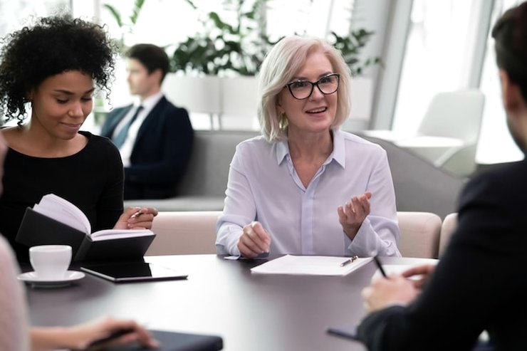 A woman with glasses speaking to a small group at a table, with notebooks and a tablet in front of them