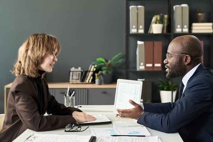 A woman and a man facing each other across a desk, with a tablet and printed papers between them